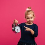 happy young blonde girl holding alarm clock pointing it